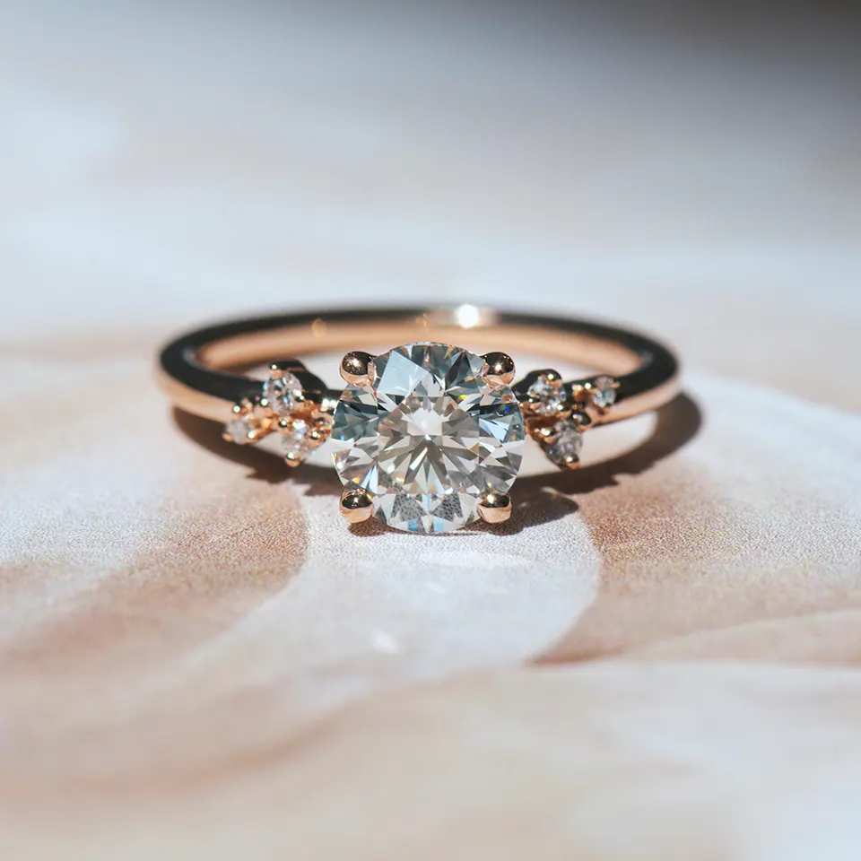 Round brilliant diamond engagement ring in rose gold with four prongs and a cluster of small diamonds on each shoulder