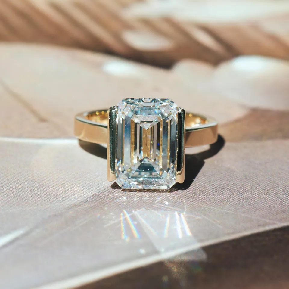 Emerald-cut diamond engagement ring in a yellow gold half-bezel on a wide flat band, in sunlight