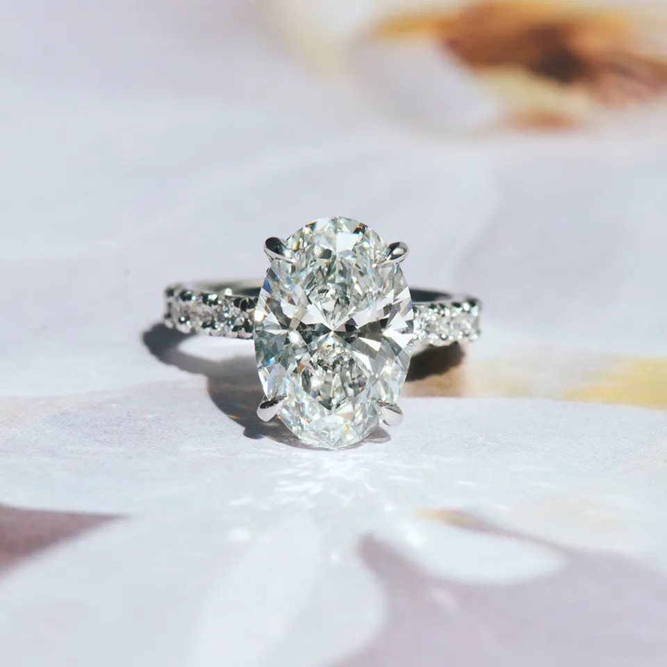 Oval diamond engagement ring in four claw prongs on a white metal band set with round diamonds, on white silk
