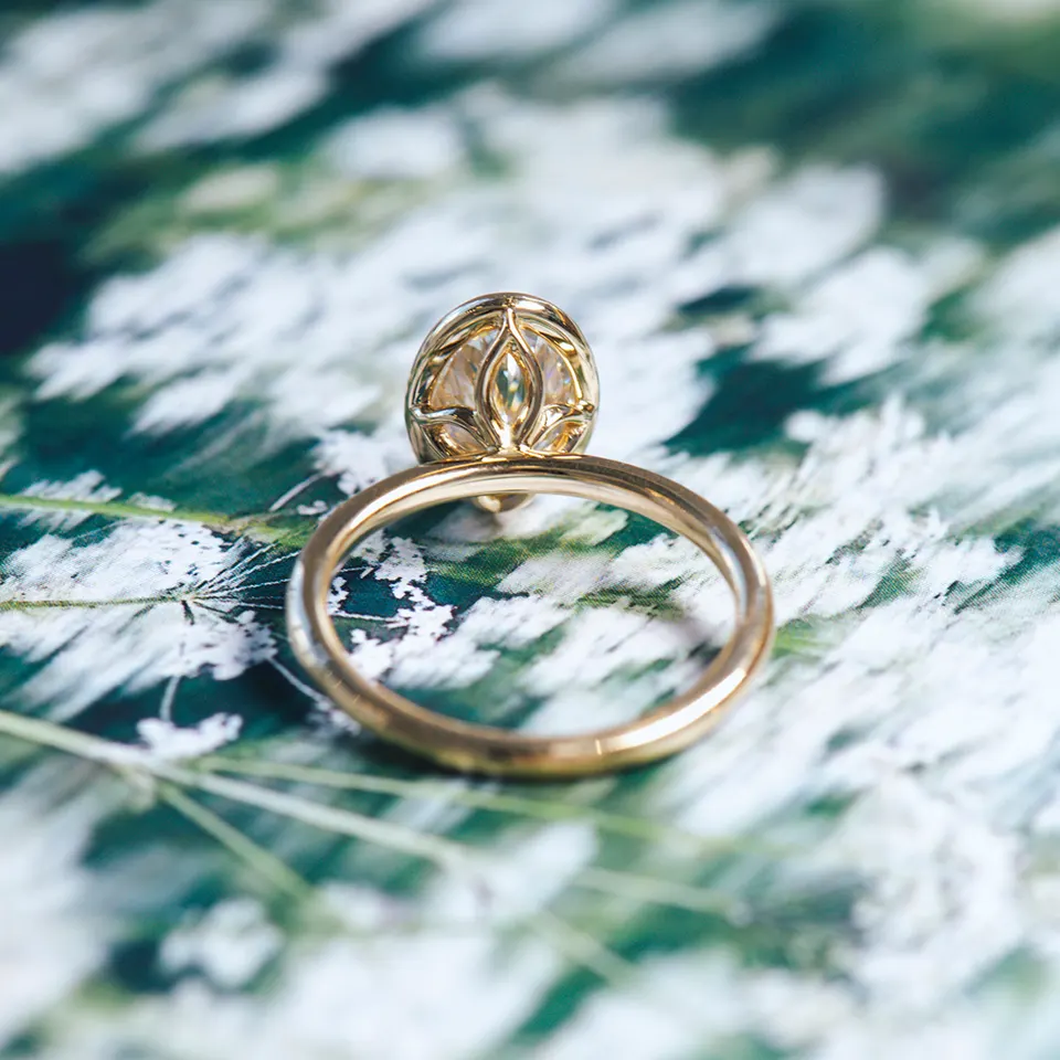 Back view of a yellow gold engagement ring showing the oval stone's lotus-pattern gallery, on a green botanical print