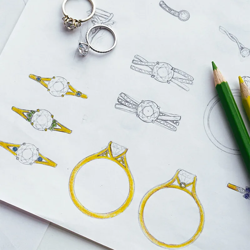 Custom engagement ring sketches in pencil and yellow, beside two finished rings and coloured pencils